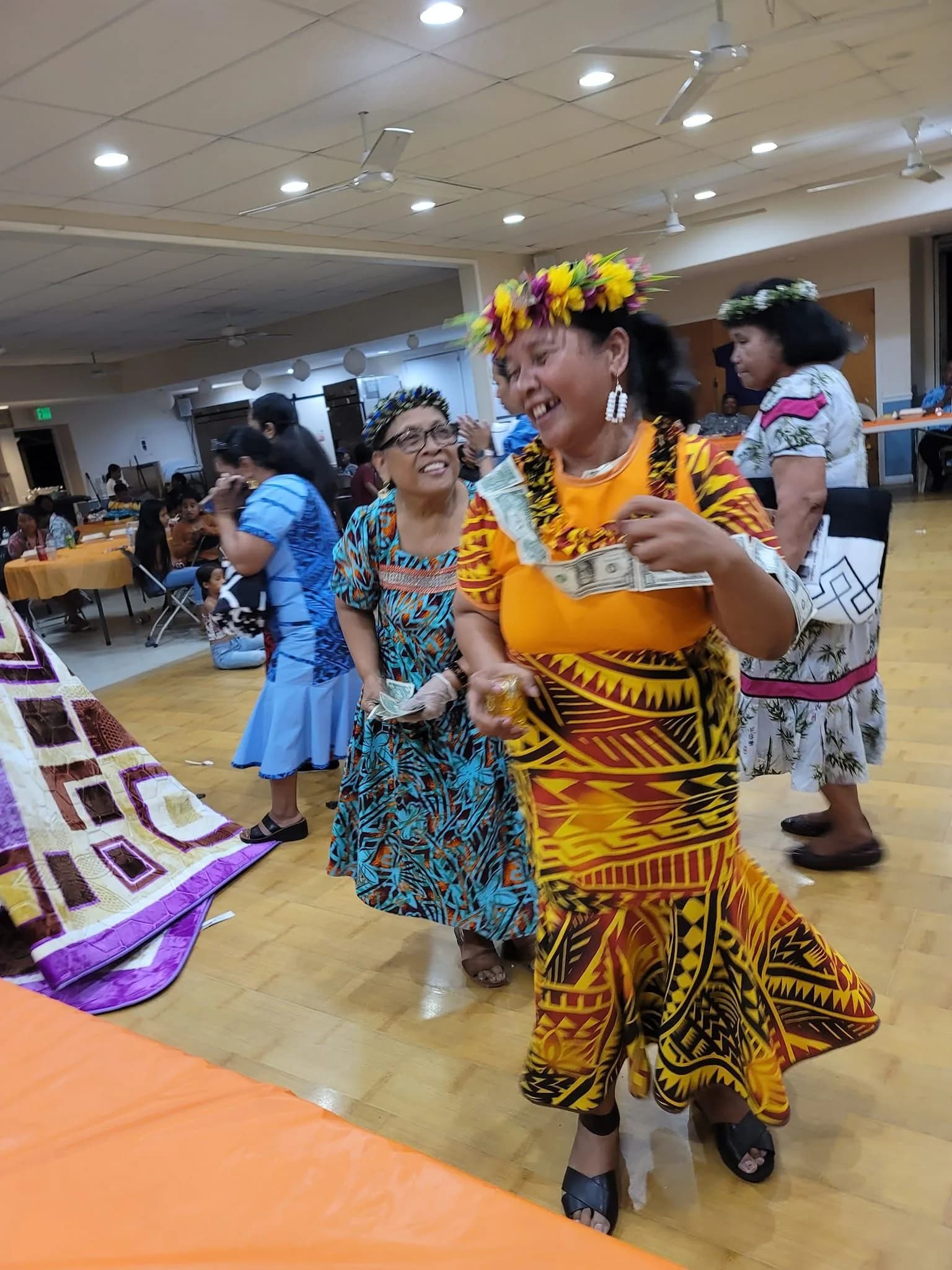 Women of the church laughing as they dance during a fellowship night, wearing flower crowns and gold and red island dress.